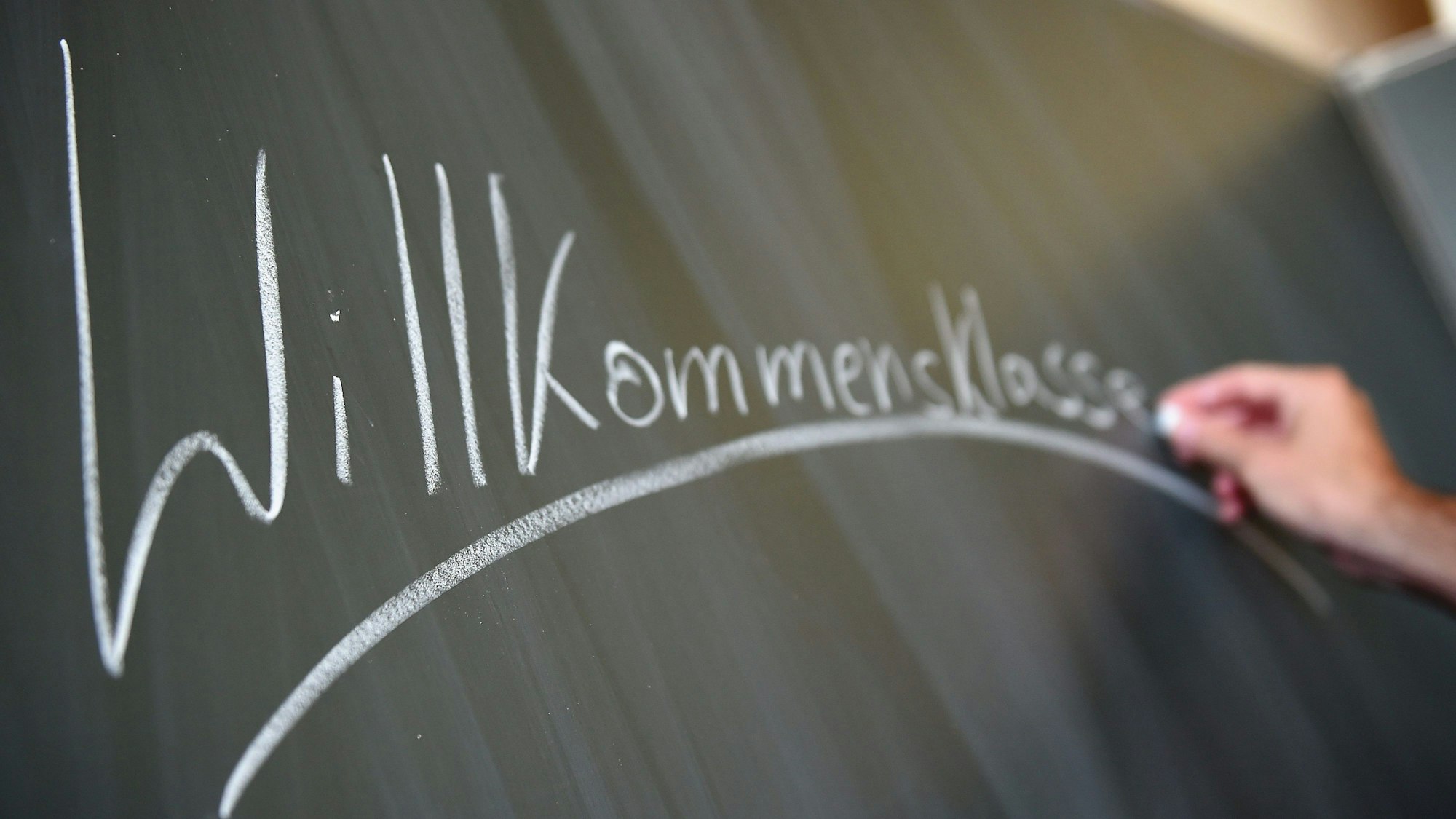 Eine Hand schreibt mit weißer Kreide das Wort „Willkommensklasse“ an eine Tafel.