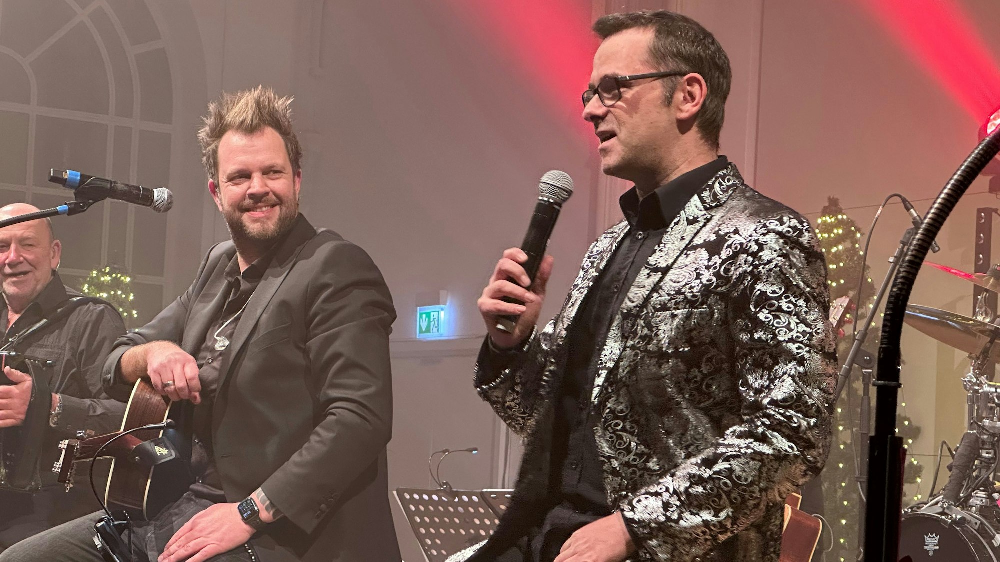 Der Kabarettist Marc Metzger sitzt im Glitzerjacket auf der Bühne der Flora bei den Weihnachtskonzerten der Paveier. Links neben ihm lauschen die Musiker Sven Welter und Detlef Vorholt.