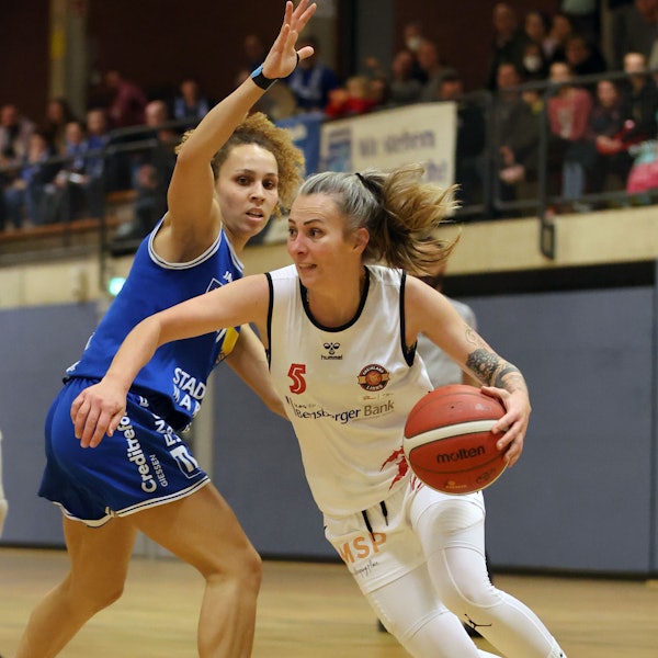 Im weißen Trikot führt die Spielerin der Rheinland Lions den Ball an der Hand. Ihre Gegnerin im blauen Trikot versucht sie daran zu hindern.