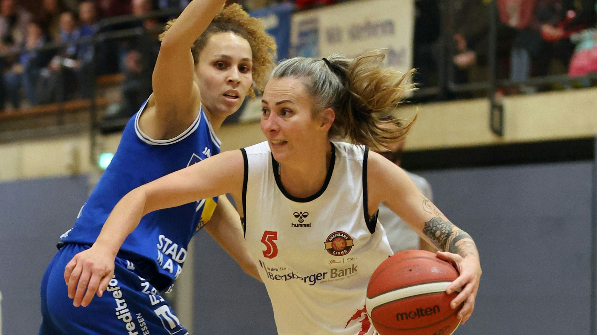 Im weißen Trikot führt die Spielerin der Rheinland Lions den Ball an der Hand. Ihre Gegnerin im blauen Trikot versucht sie daran zu hindern.