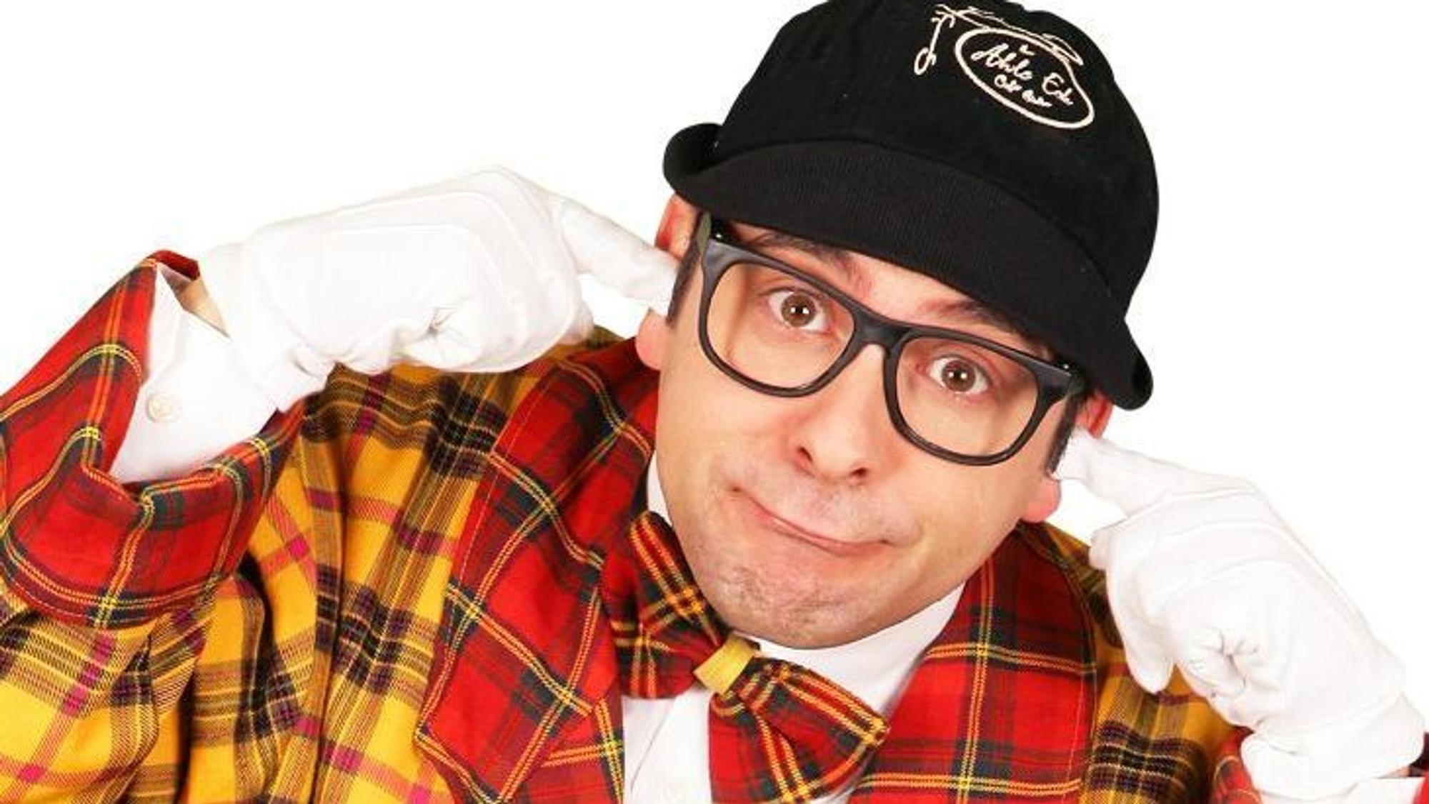 Der Komiker Marc Metzger trägt einen rot-gelb karierten Anzug mit Fliege, Kappe und Brille, guckt verdötscht und steckt sich die weiß behandschuhten Zeigefinger in die Ohren