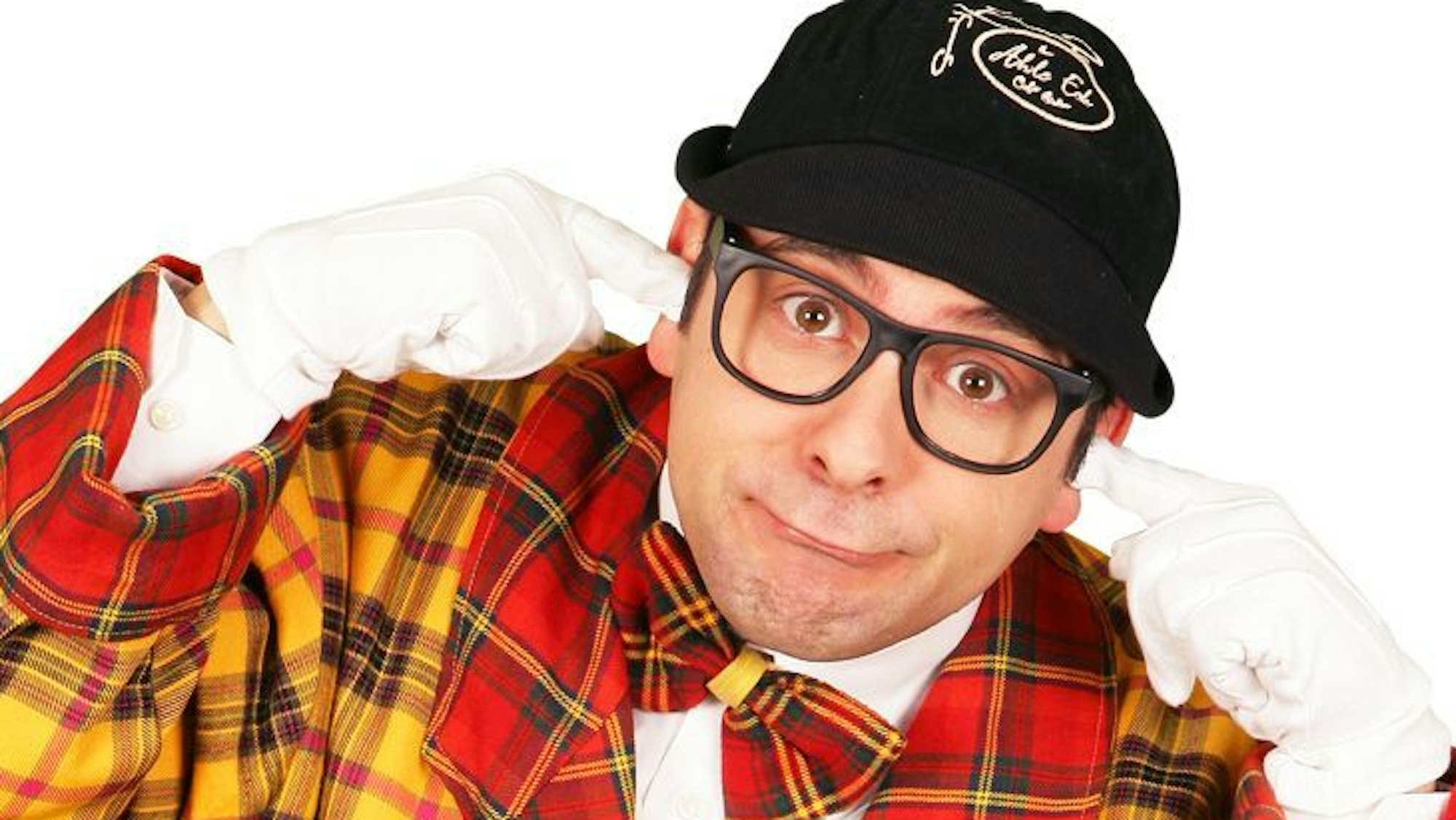 Der Komiker Marc Metzger trägt einen rot-gelb karierten Anzug mit Fliege, Kappe und Brille, guckt verdötscht und steckt sich die weiß behandschuhten Zeigefinger in die Ohren