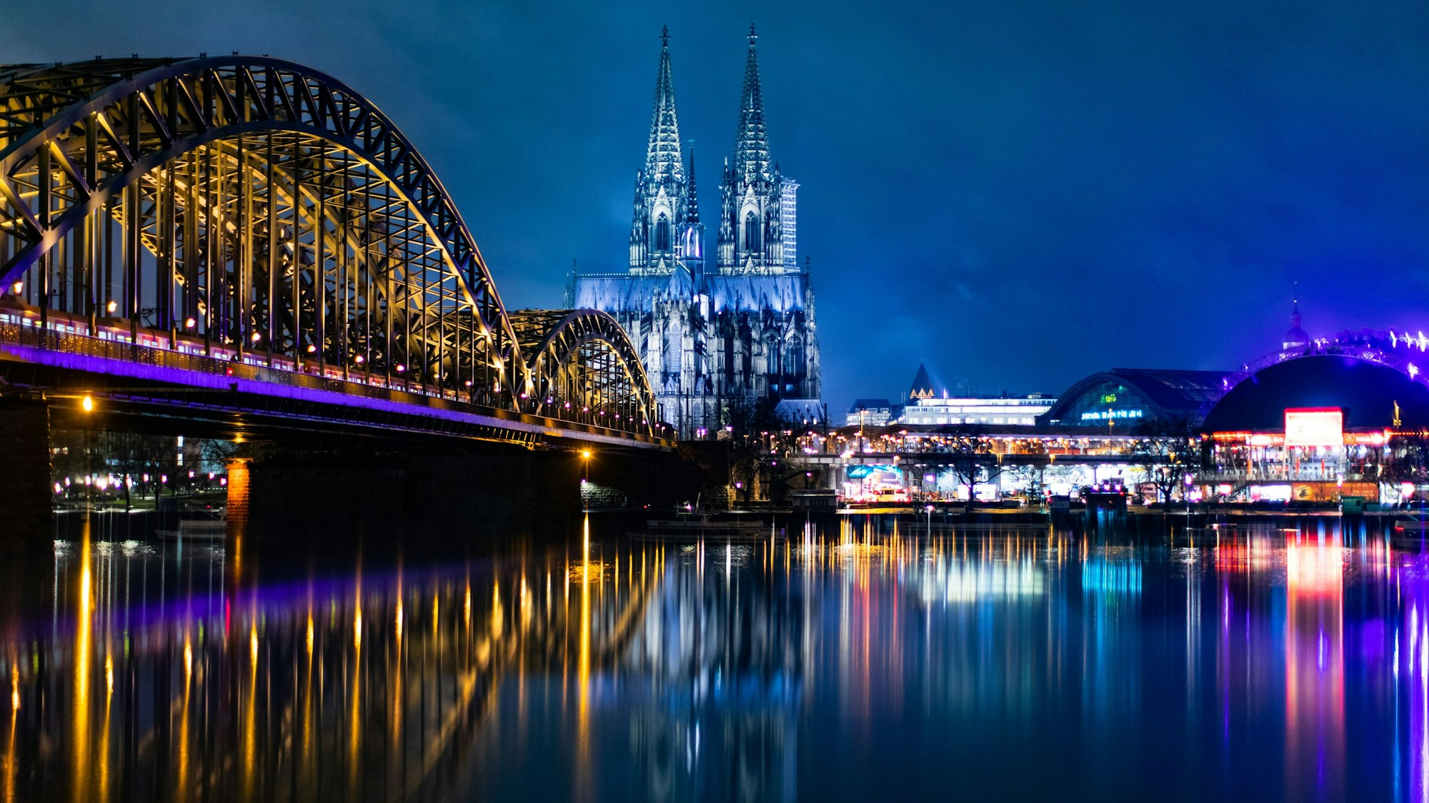 Die bunt beleuchtete Hohenzollernbrücke mit dem Kölner Dom und dem Musical Dome bei Nacht.