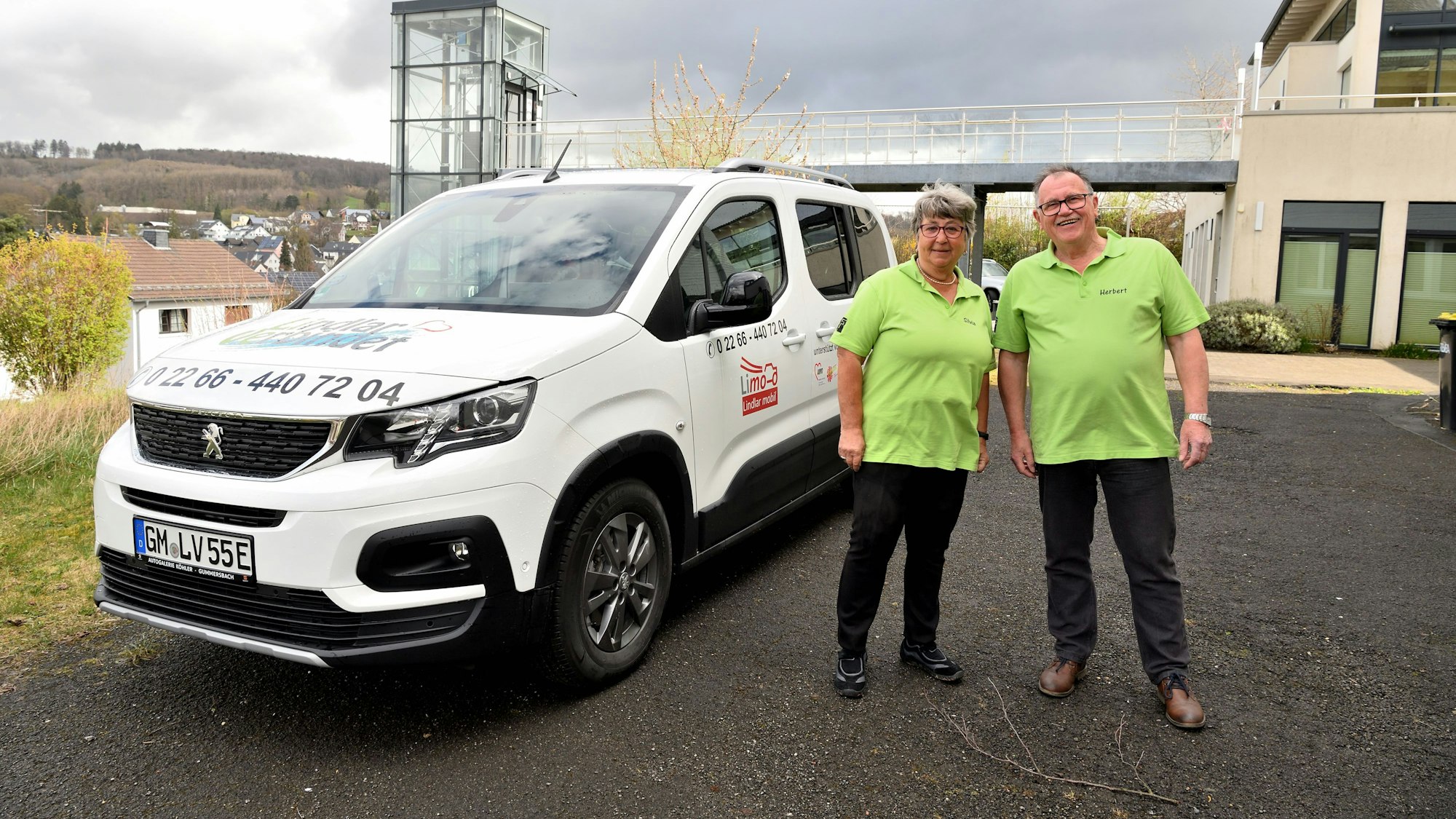 Silvia Siebenmorgen und Herbert Schmitz stehen in grünen T-Shirts vor einem weißen Fahrzeug mit den Kontaktdaten des Fahrdienstes.