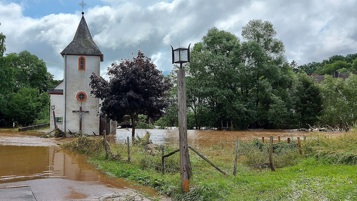 Hochwasser bedeckt eine Straße. Ein Haus und eine Kirche stehen im Wasser. Nur ein wenig Wiese kommt an der Seite des Bildes noch aus dem Wasser.