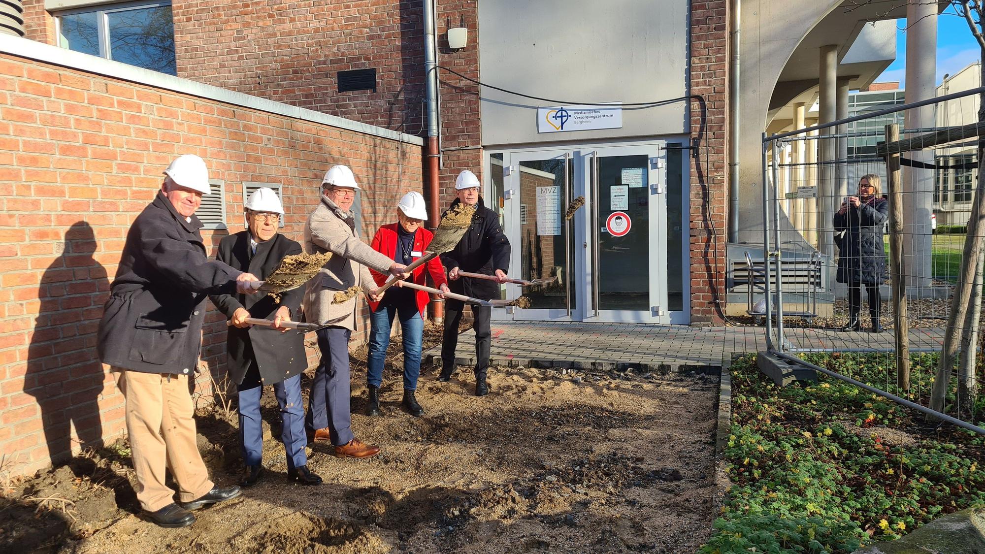 Fünf Männer mit Bauhelmen stehen vor dem Krankenhaus auf Baustellenboden und haben einen Spaten i der Hand.