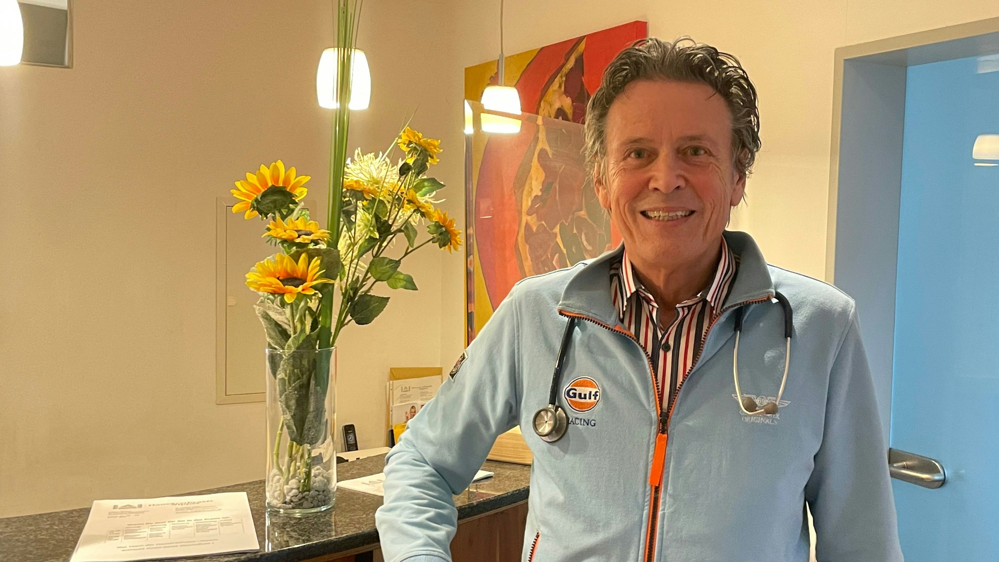 Hausarzt Roland Wollny steht in seiner Praxis in Köln-Nippes und blickt lächelnd in die Kamera.