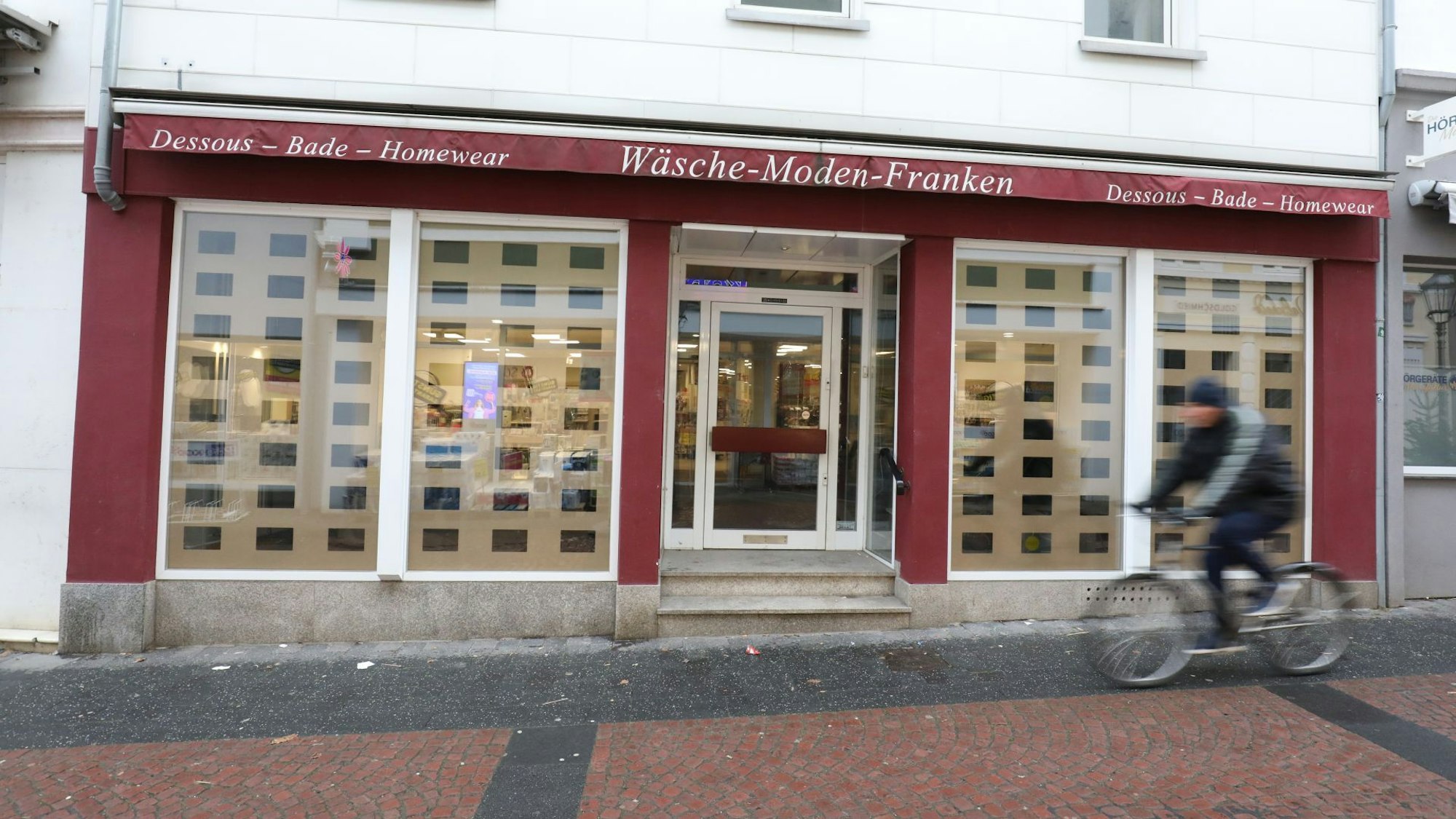 An einem leeren Geschäft mit zum Teil roter Fassade fährt ein Radfahrer vorbei.