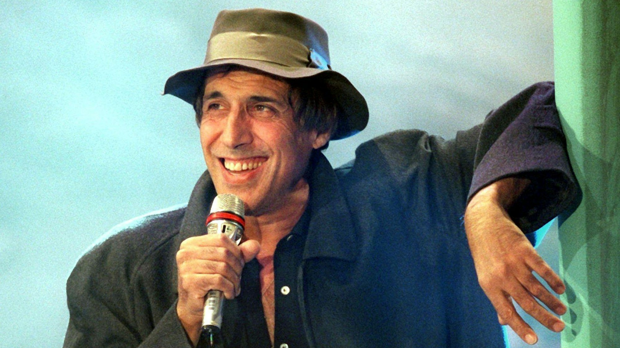 Adriano Celentano lehnt sich mit dem linken Arm an, in der rechten Hand hält er ein Mikrofon und lächelt. Er trägt einen Hut.