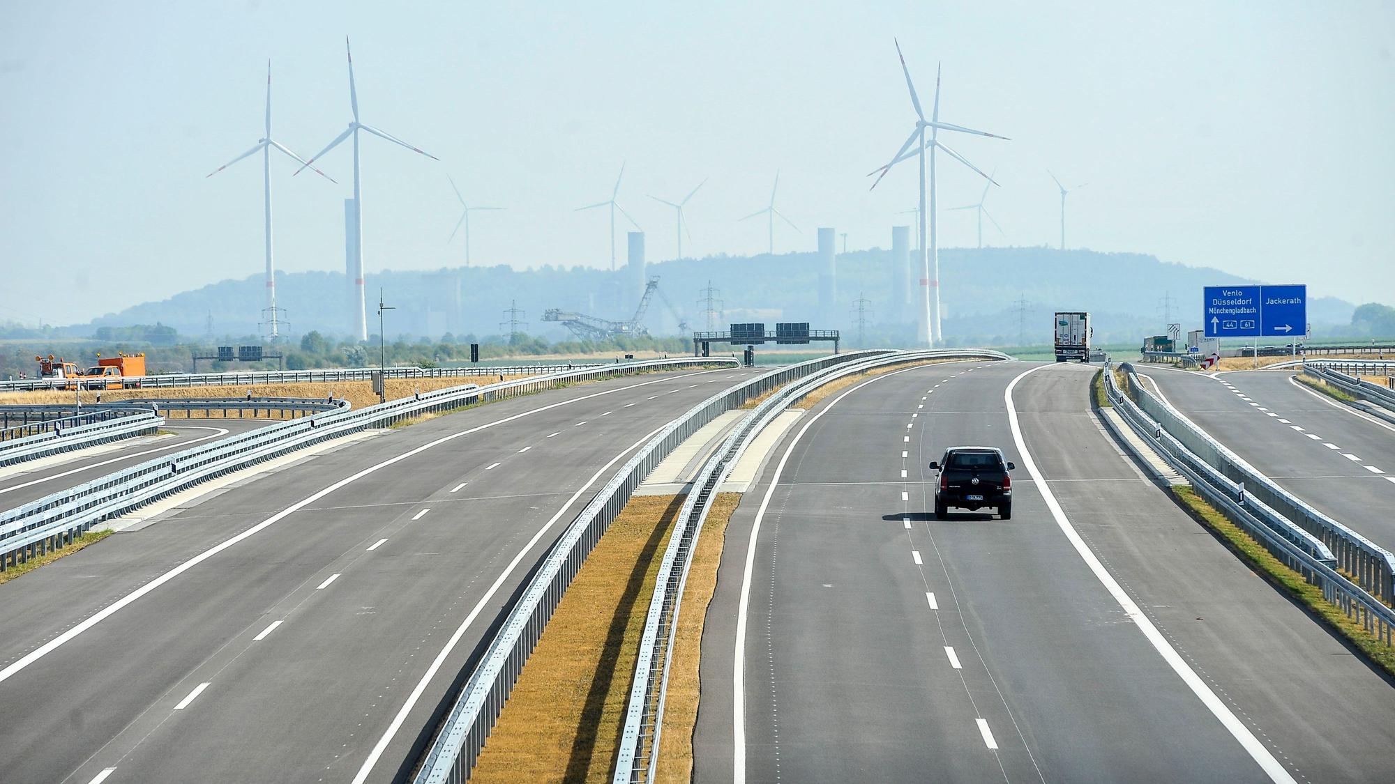 Ein Auto fährt auf einer Autobahn. Im Hintergrund sind Windräder und der obere Teil eines Braunkohle-Baggers zu sehen.