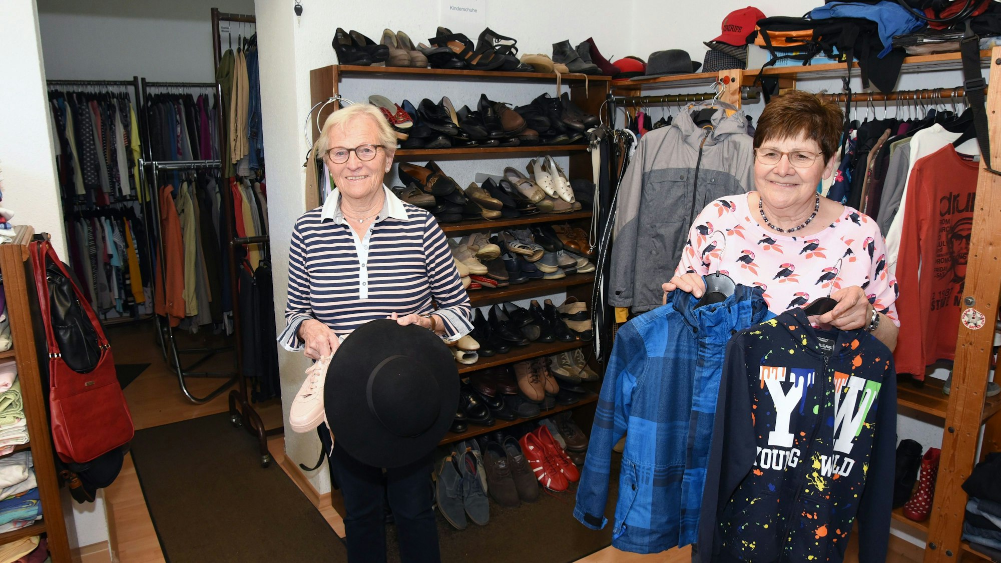 Adeline Zanders (l.) und Agnes Ahlers stehen in ihrem Laden und halten Klamotten – eine Jacke, einen Pulli, einen Hut und Schuhe.