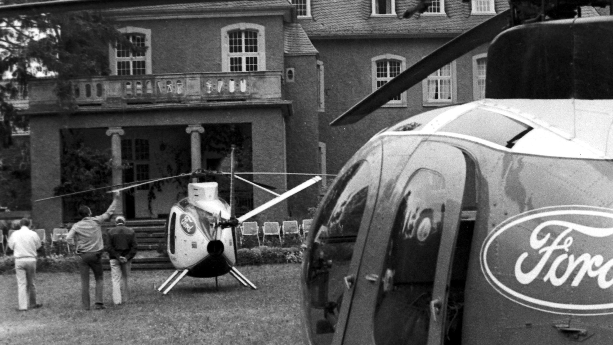 Zwei Hubschrauber mit Ford-Logos steht vor der Burg Dalbenden, daneben drei von der Kamera abgewandte Personen. Das Foto ist in schwarz-weiß.