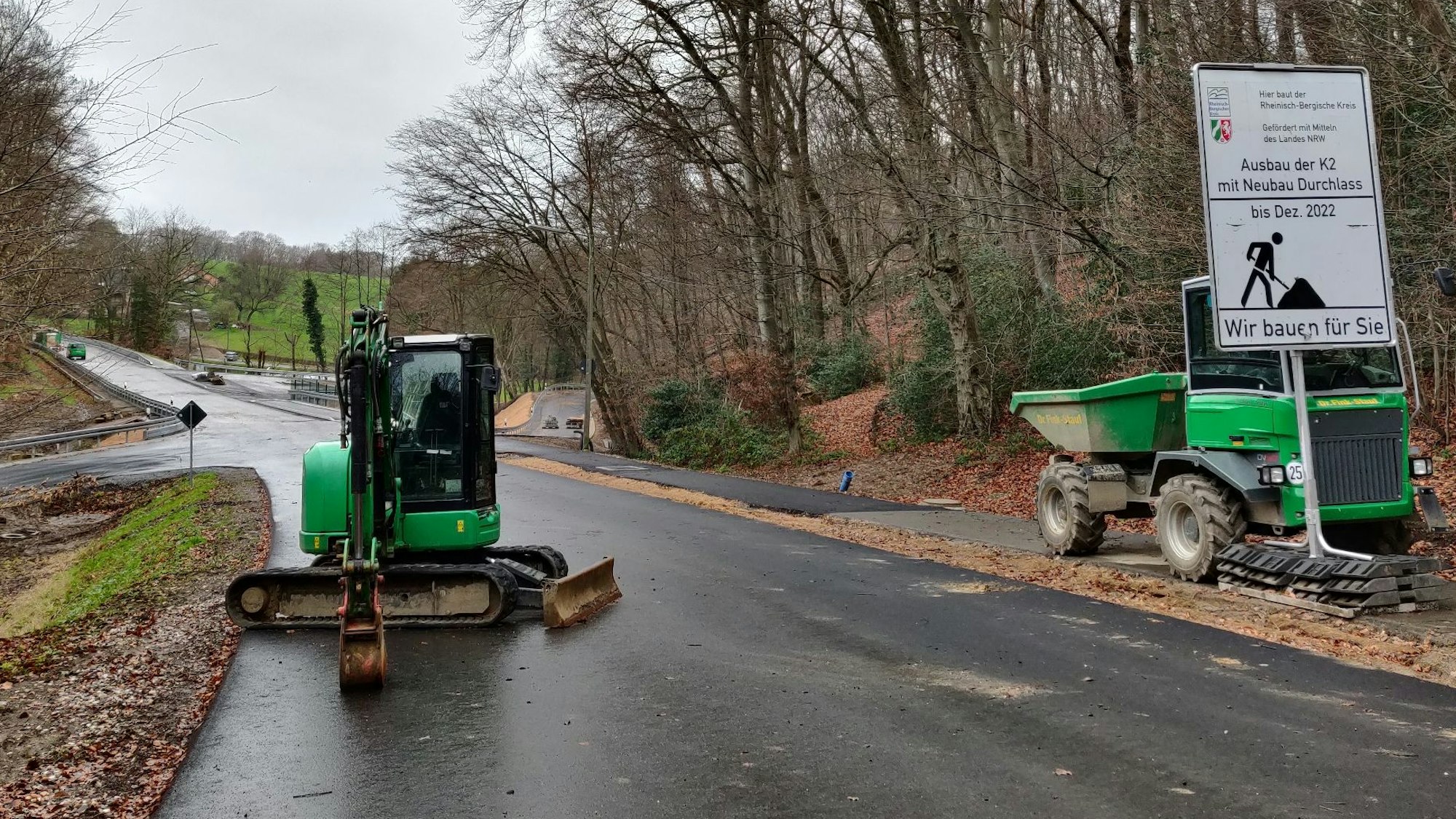 Auf einem Baustellenschild neben der Straße bei Dohm wird das Ende der Baustelle für Dezember 2022 angekündigt. Daneben parken ein grüner Muldenkipper und ein Raupenbagger.