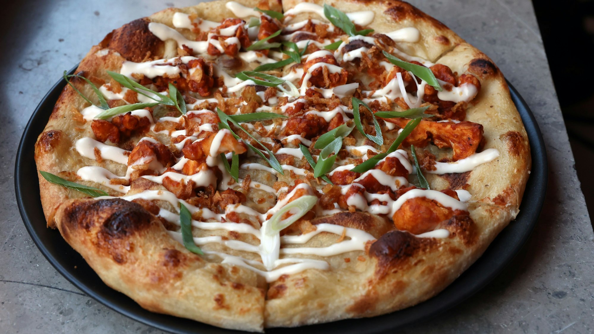 Eine vegane Pizza des Restaurants „Virtuous Pie“ liegt verzehrbereit auf einem schwarzen Teller.
