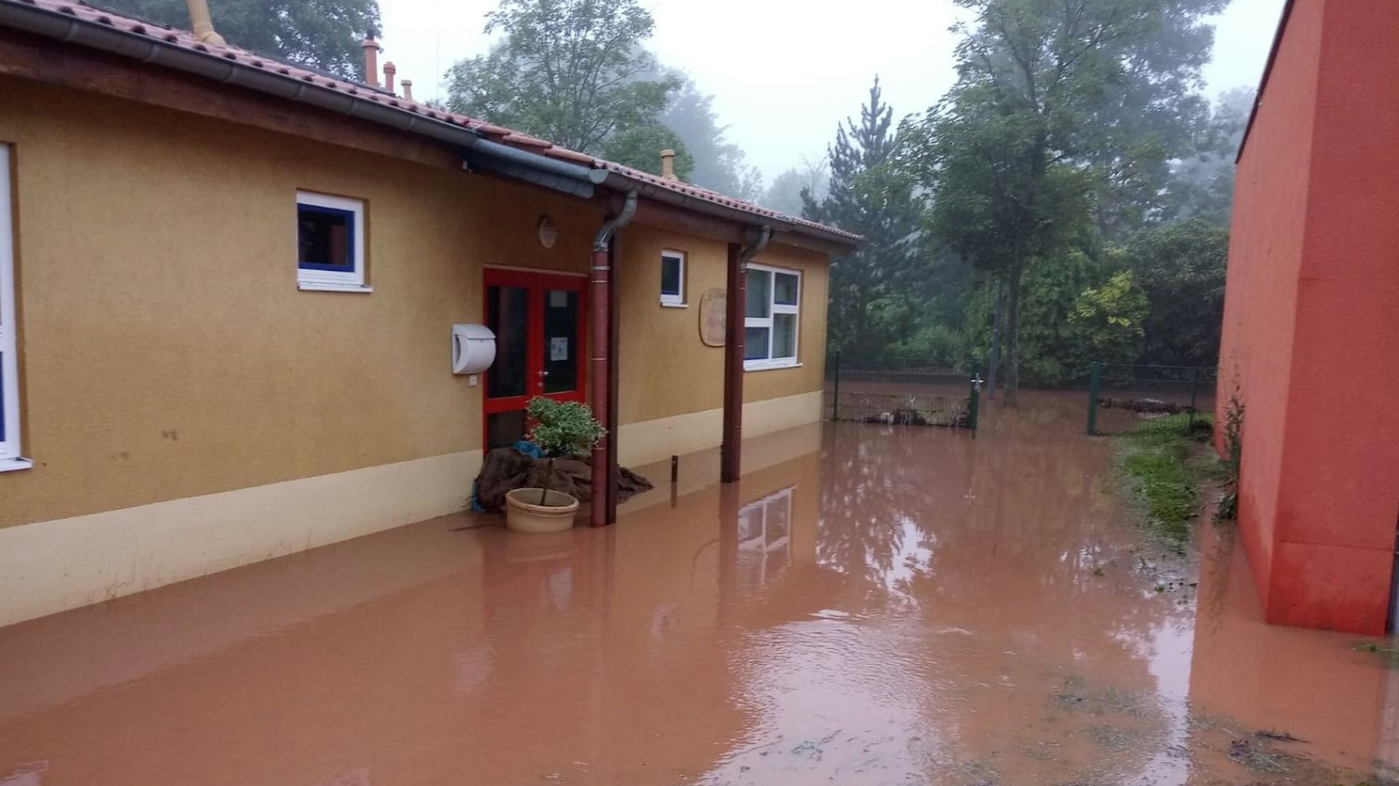 Ein ockerfarbenes Gebäude ist umgeben von rot-braunem Wasser. Im Hintergrund sind Bäume zu erkennen.