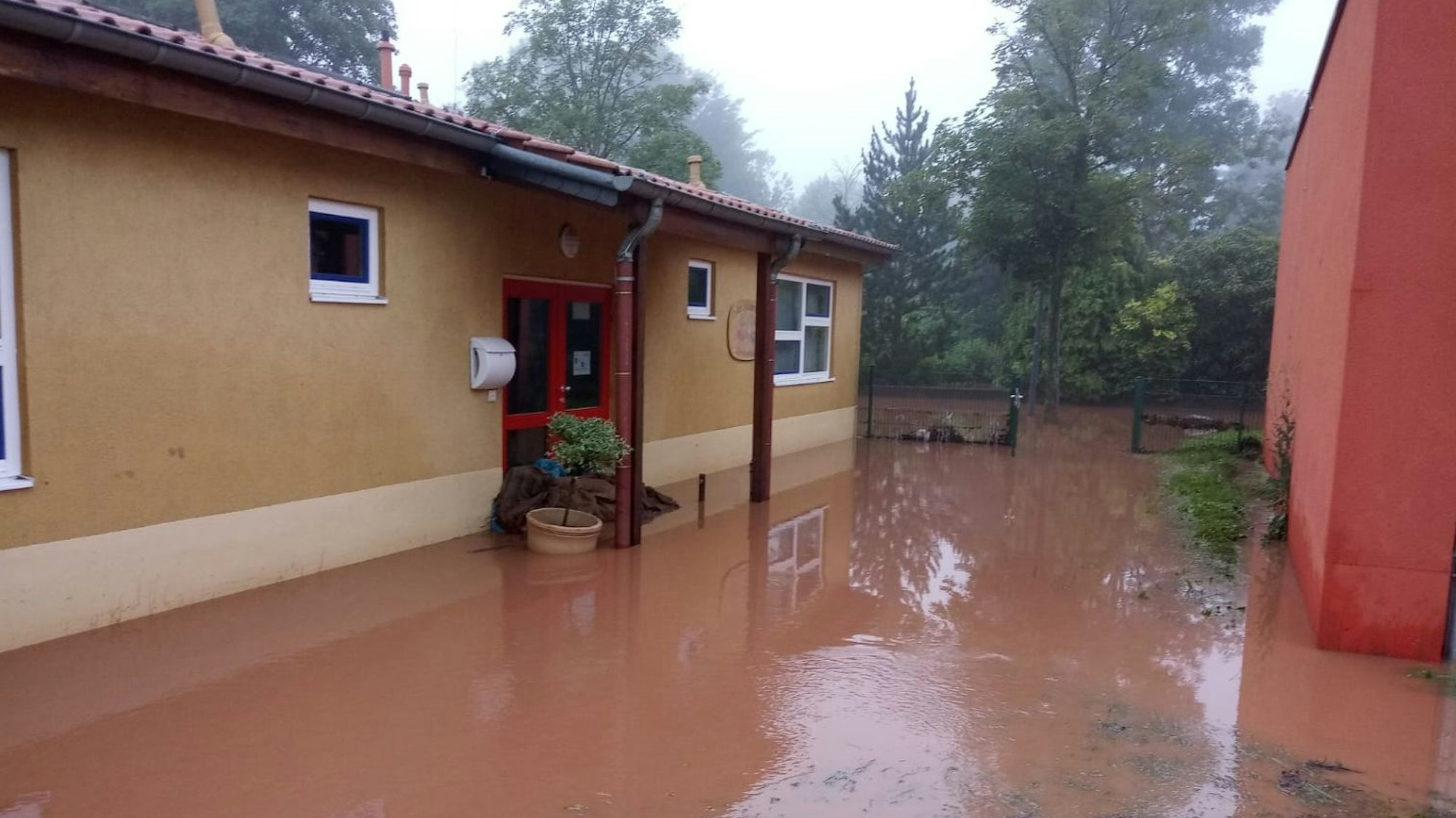 Ein ockerfarbenes Gebäude ist umgeben von rot-braunem Wasser. Im Hintergrund sind Bäume zu erkennen.