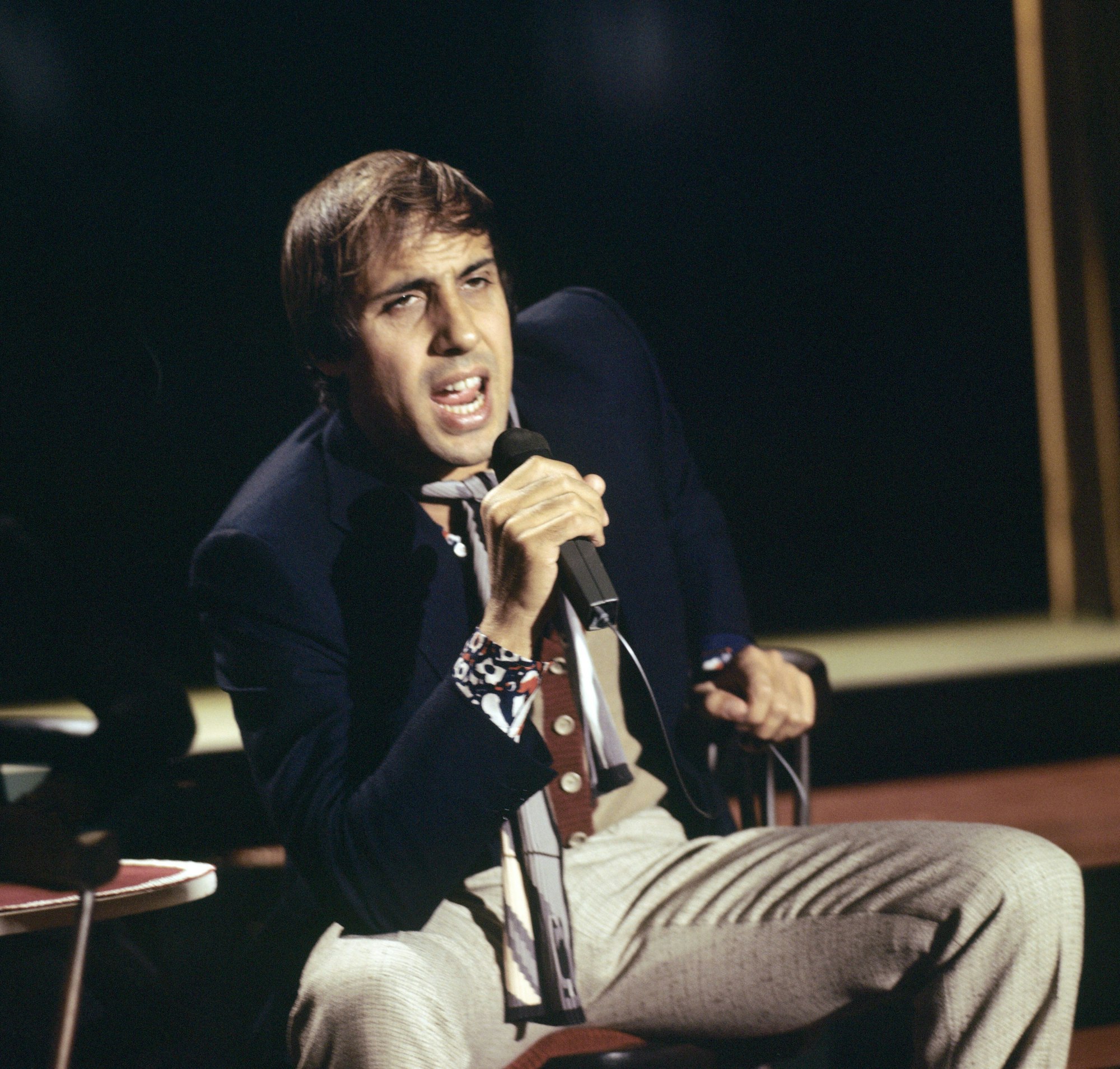 Adriano Celentano sitzt auf einem Stuhl und singt in ein Mikrofon. Er trägt eine helle Hose und ein dunkelblaues Sakko.