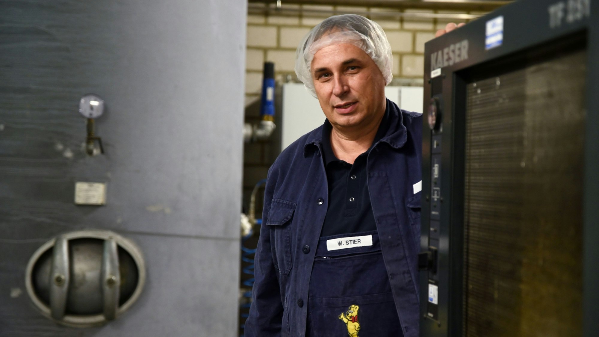 Wolfgang Stier trägt blaue Arbeitskleidung und eine durchsichtige Haube über dem Kopf. Er steht in einer Fabrik.