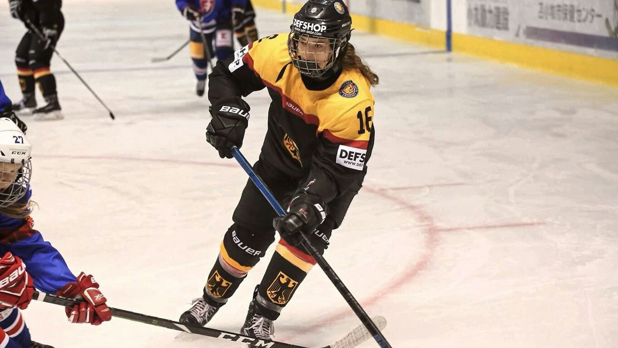 Lea Badura steht in voller Eishockey-Montur und mit Schläger auf dem Eis. Sie fährt gerade in Richtung Kamera, neben ihr fährt eine Spielerin vom gegnerischen Team.