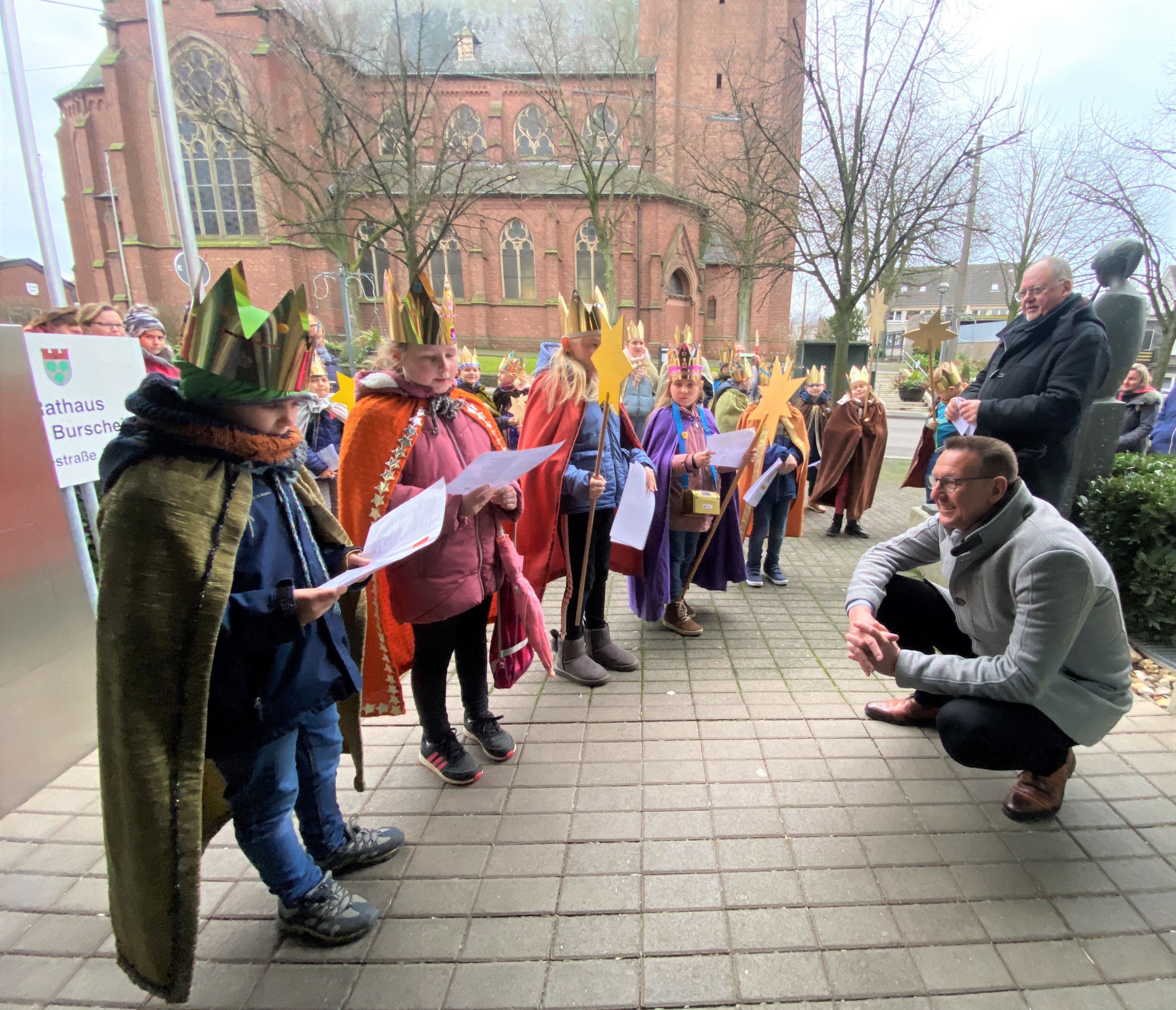Als Sternsinger verkleidete Kinder stehen auf der linken Bildseite und singen ein Lied. Bürgermeister Dirk Runge hockt auf der rechten Seite.