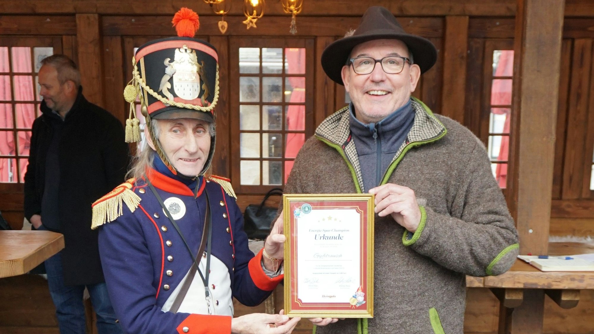 Marktgendarm Gilbert überreicht Händler Mario Michalak die Urkunde als Sieger des Energiespar-Wettbewerbs. Die beiden stehen vor einer Bude des Weihnachtsmarkts aus Holz.