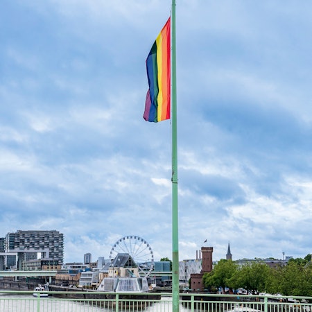 Auf der Deutzer Brücke hängt eine Regenbogenflagge. Im Hintergrund die Kranhäuser und ein Riesenrad. Das Bild stammt aus dem Pride Month im Juni 2022.