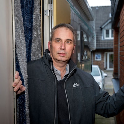 Wolfgang Stier steht zwischen zwei Häusermauern und hat seine linke Hand an den Backstein gelegt. Er trägt eine dunkle Adidas-Jacke, darunter ein Hemd und schwarzen Pullover. Er hat kurze, graue Haare.