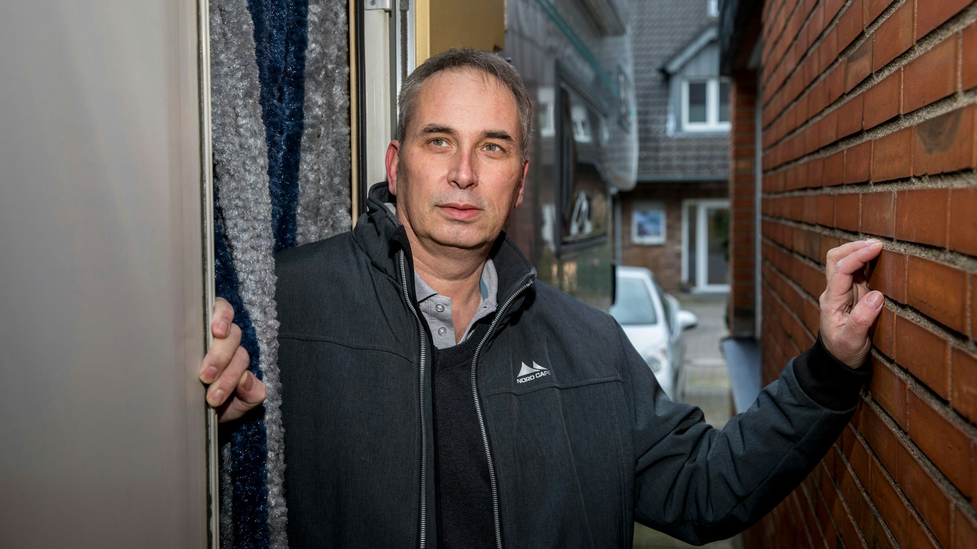 Wolfgang Stier steht zwischen zwei Häusermauern und hat seine linke Hand an den Backstein gelegt. Er trägt eine dunkle Adidas-Jacke, darunter ein Hemd und schwarzen Pullover. Er hat kurze, graue Haare.