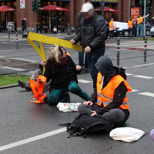 Aktivistinnen der „Letzten Generation“ sitzen auf der Fahrbahn der Cäcilienstraße in Köln. Ein Passant versucht, die Aktion zu unterbinden.