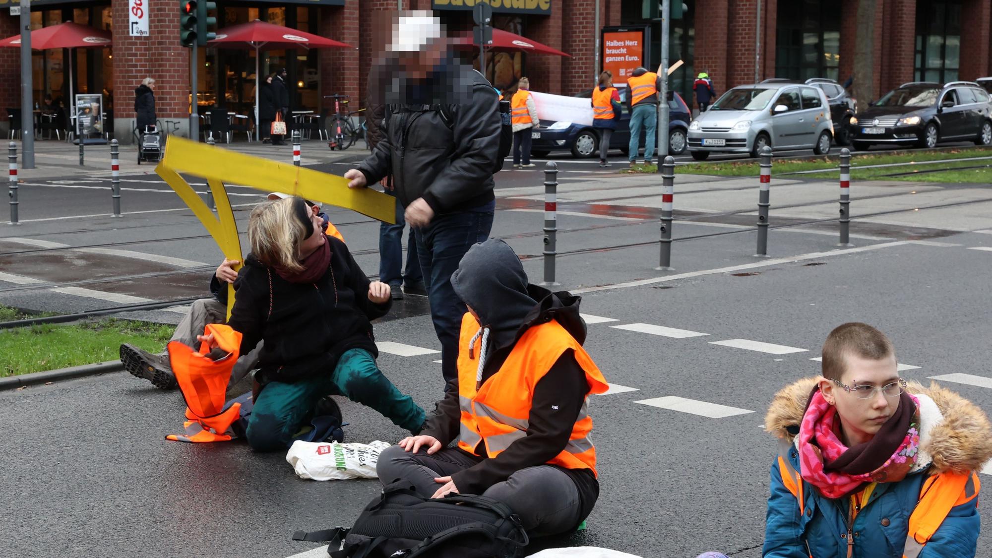 Aktivistinnen der „Letzten Generation“ sitzen auf der Fahrbahn der Cäcilienstraße in Köln. Ein Passant versucht, die Aktion zu unterbinden.