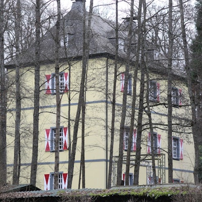 Die Burg Welterode in Eitorf mit gelber Fassade und weiß-roten Fensterläden, umringt von Bäumen.