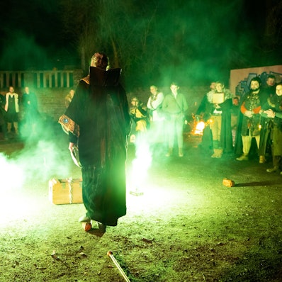 Ein Mann in Magierkostüm steht vor einer Kiste, neben ihm steigen grüne Feuerfontänen und Rauch empor.