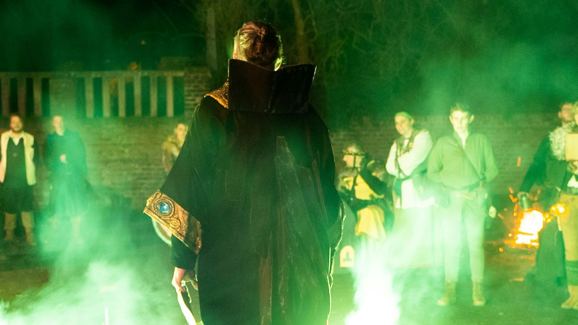 Ein Mann in Magierkostüm steht vor einer Kiste, neben ihm steigen grüne Feuerfontänen und Rauch empor.