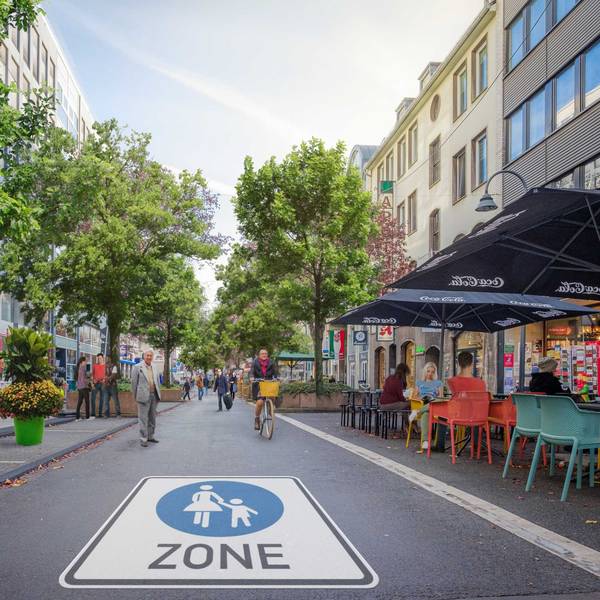 Auf der Straße prangt ein riesiges Piktogramm einer Fußgängerzone. Es sind Radfahrer und Fußgänger unterwegs, in einem Straßencafé sitzen Menschen.