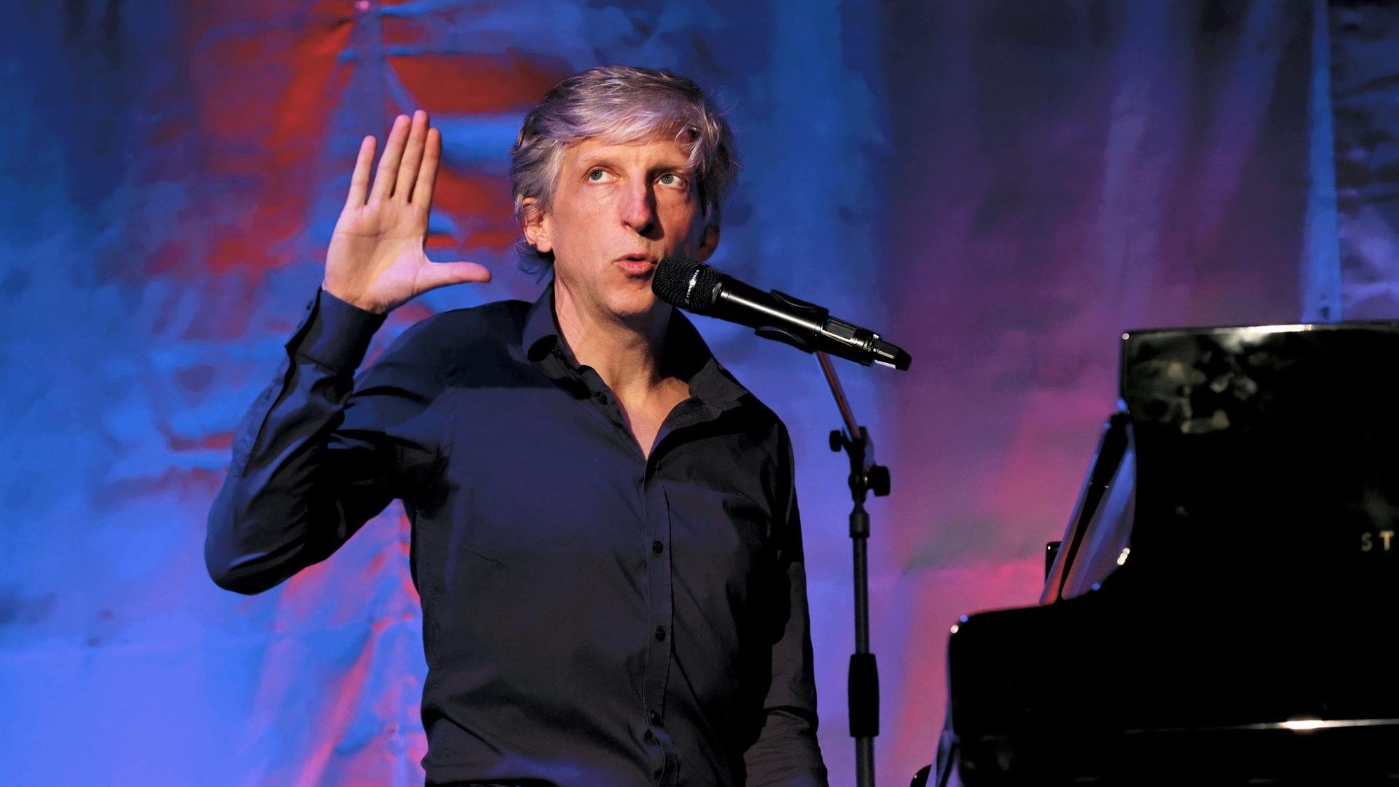 Der Kabarettist William Wahl sitzt an einem schwarzen Piano, macht eine Handgeste und singt.