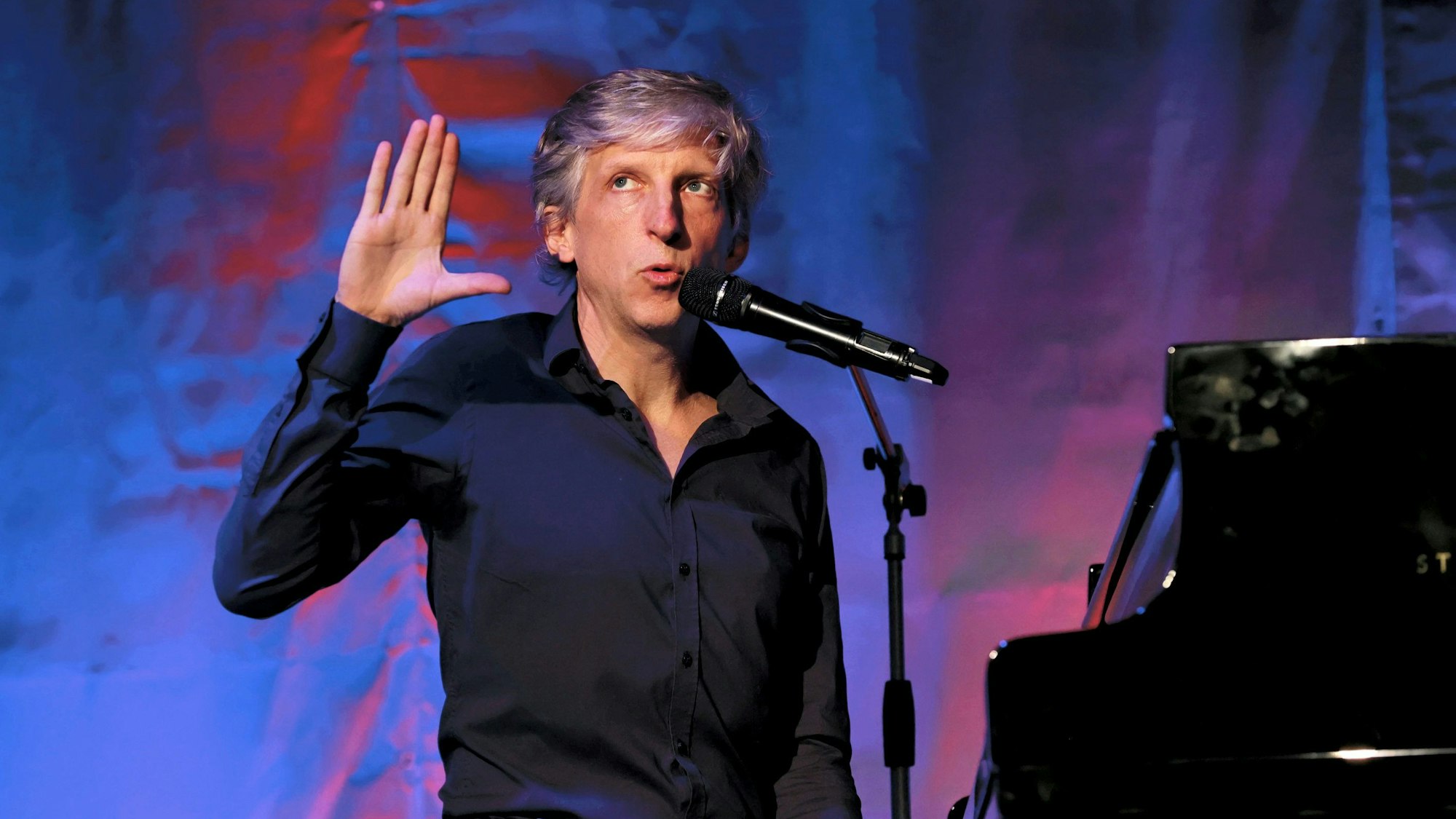 Der Kabarettist William Wahl sitzt an einem schwarzen Piano, macht eine Handgeste und singt.