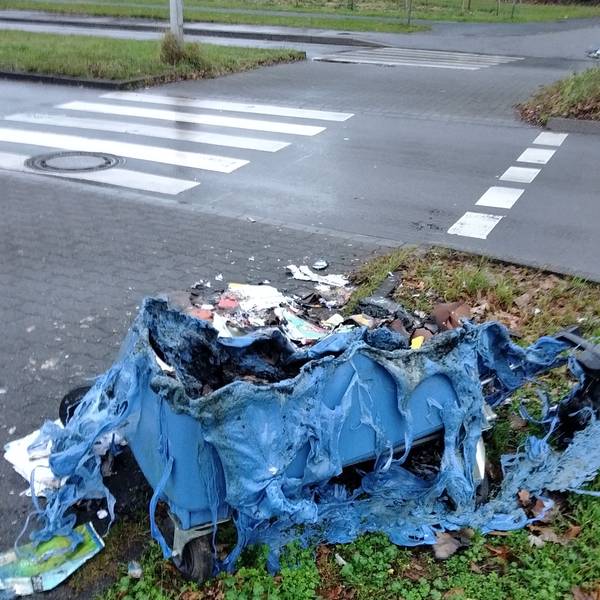 Ausgebrannte Altpapiercontainer liegen am Straßenrand vor einem Kreisverkehr.