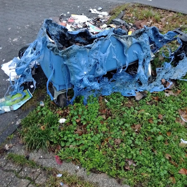 Ausgebrannte Altpapiercontainer liegen am Straßenrand vor einem Kreisverkehr.