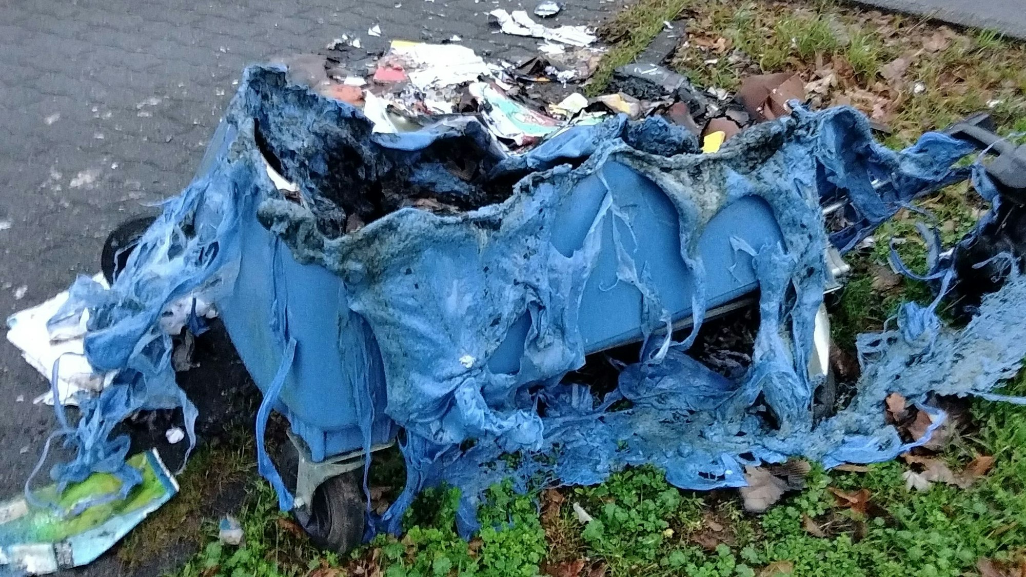 Ausgebrannte Altpapiercontainer liegen am Straßenrand vor einem Kreisverkehr.