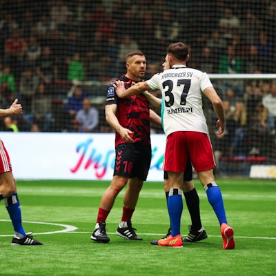 Lukas Podolski wird von einem Mann im weißen Trikot an der Schulter gefasst. Er schaut wütend.