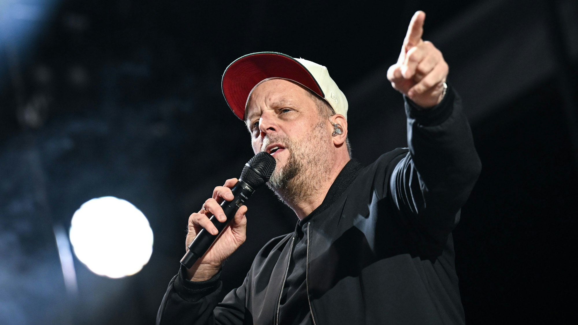 Smudo steht mit einem Mikrofon auf der Bühne, er zeigt auf etwas. Er trägt eine weiße Mütze.