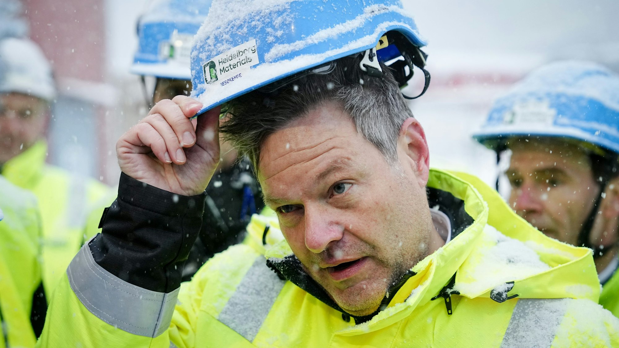 Robert Habeck (Bündnis 90/Die Grünen), Bundesminister für Wirtschaft und Klimaschutz, hebt einen blauen Bauarbeiterhelm voller Schnee nach oben. Er trägt eine neongelbe Jacke.