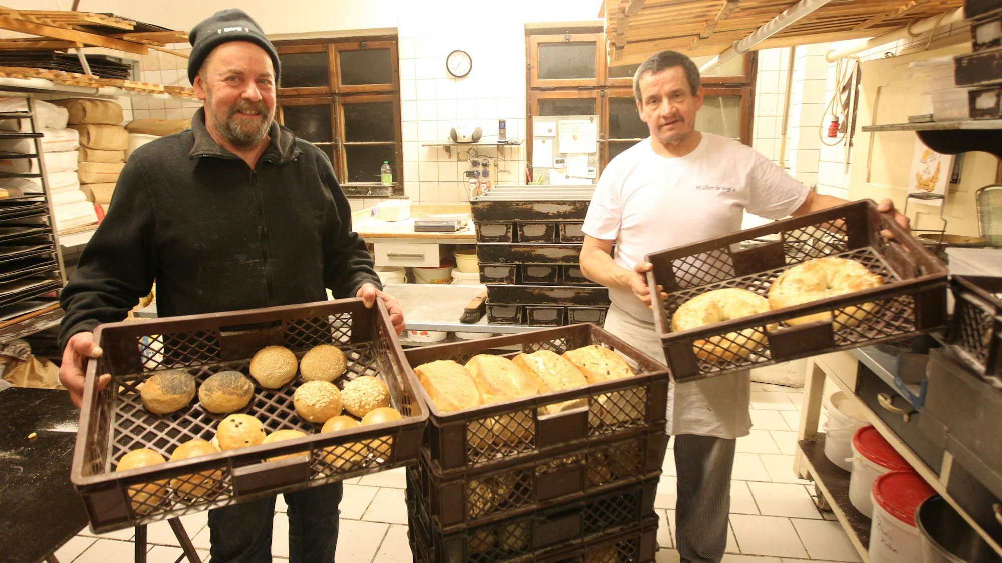 Zwei Männer stehen in einer Backstube und halten Körbe mit Broten und Brötchen in den Händen.