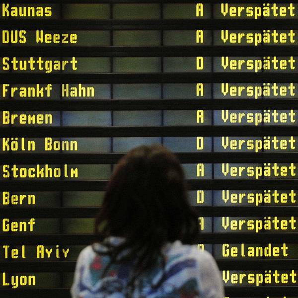 Eine Frau steht am Flughafen vor einer Anzeigetafel. Zahlreiche Flüge sind als "Verspätet" markiert.