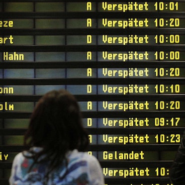 Eine Frau steht am Flughafen vor einer Anzeigetafel. Zahlreiche Flüge sind als "Verspätet" markiert.