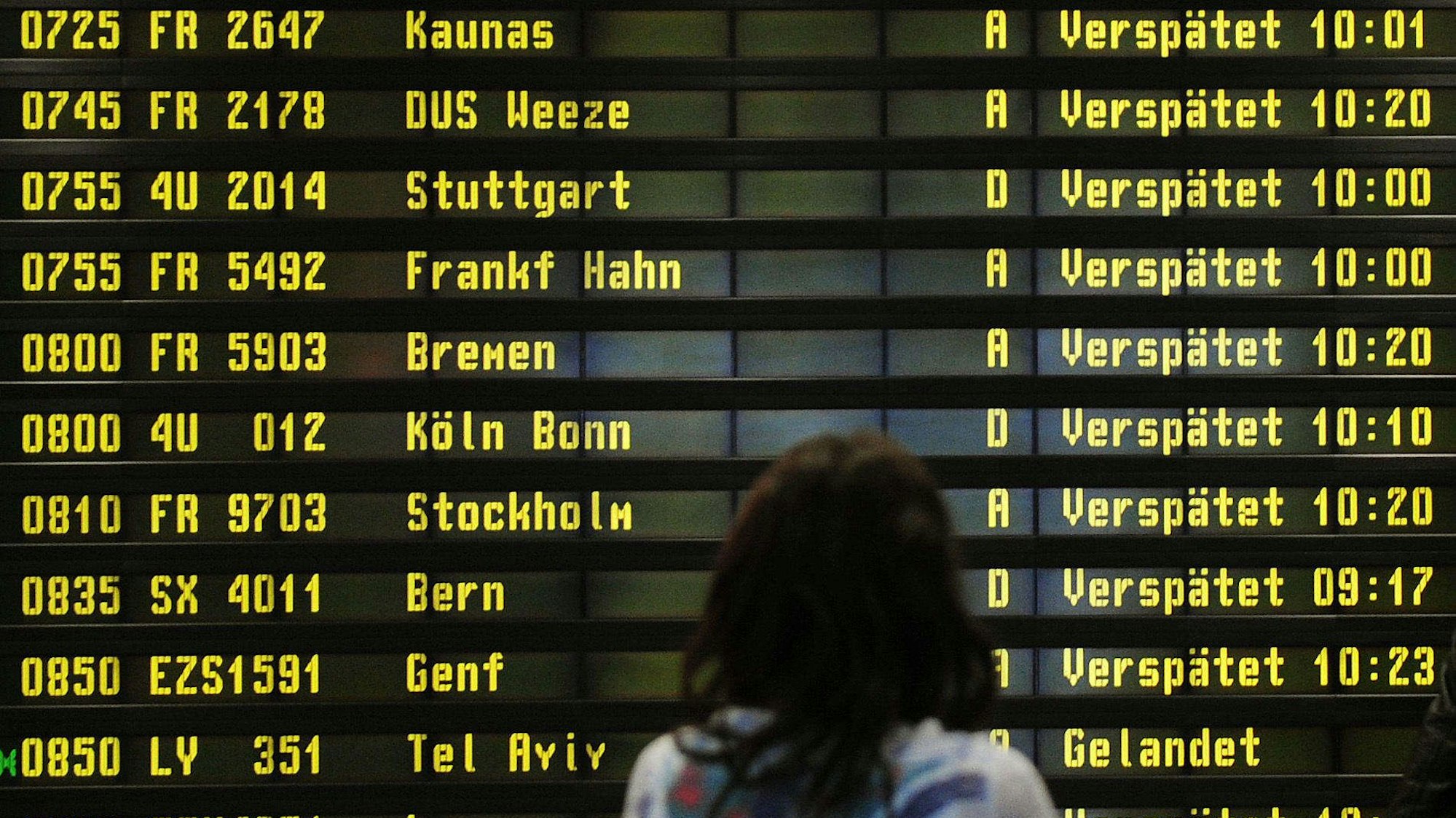 Eine Frau steht am Flughafen vor einer Anzeigetafel. Zahlreiche Flüge sind als "Verspätet" markiert.