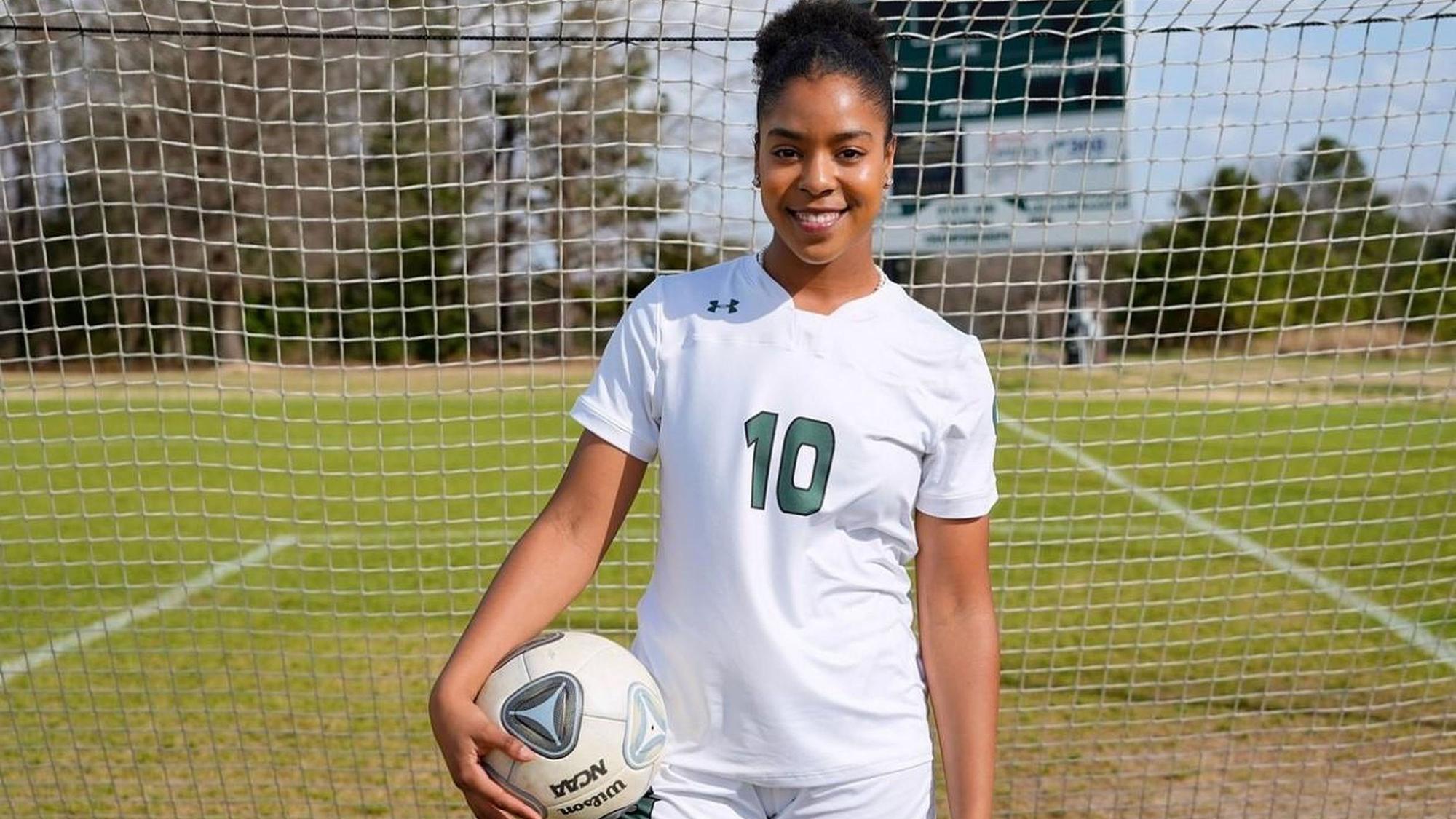 Die Fußballerin Zahra Fepessi trägt die Nummer 10 auf ihrem Trikot und steht in Fußballkleidung und mit einem Ball unter dem Arm vor einem Fußballtor.