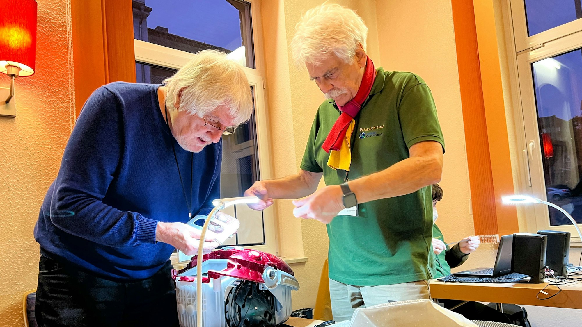 Zwei ältere Männer stehen über einem demontierten Staubsauger und schauen sich das Gerät genau an.