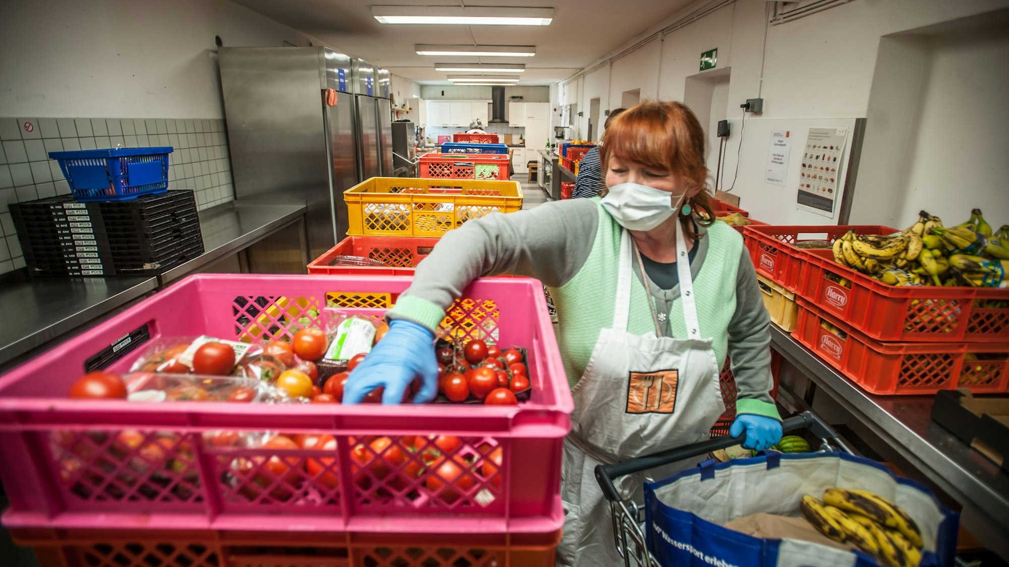 Eine Frau greift in eine Kiste voller Tomaten. In der großen Industrieküche sind weitere Körbe mit Gemüse und Obst zu erkennen, in einer sind Bananen zu sehen.