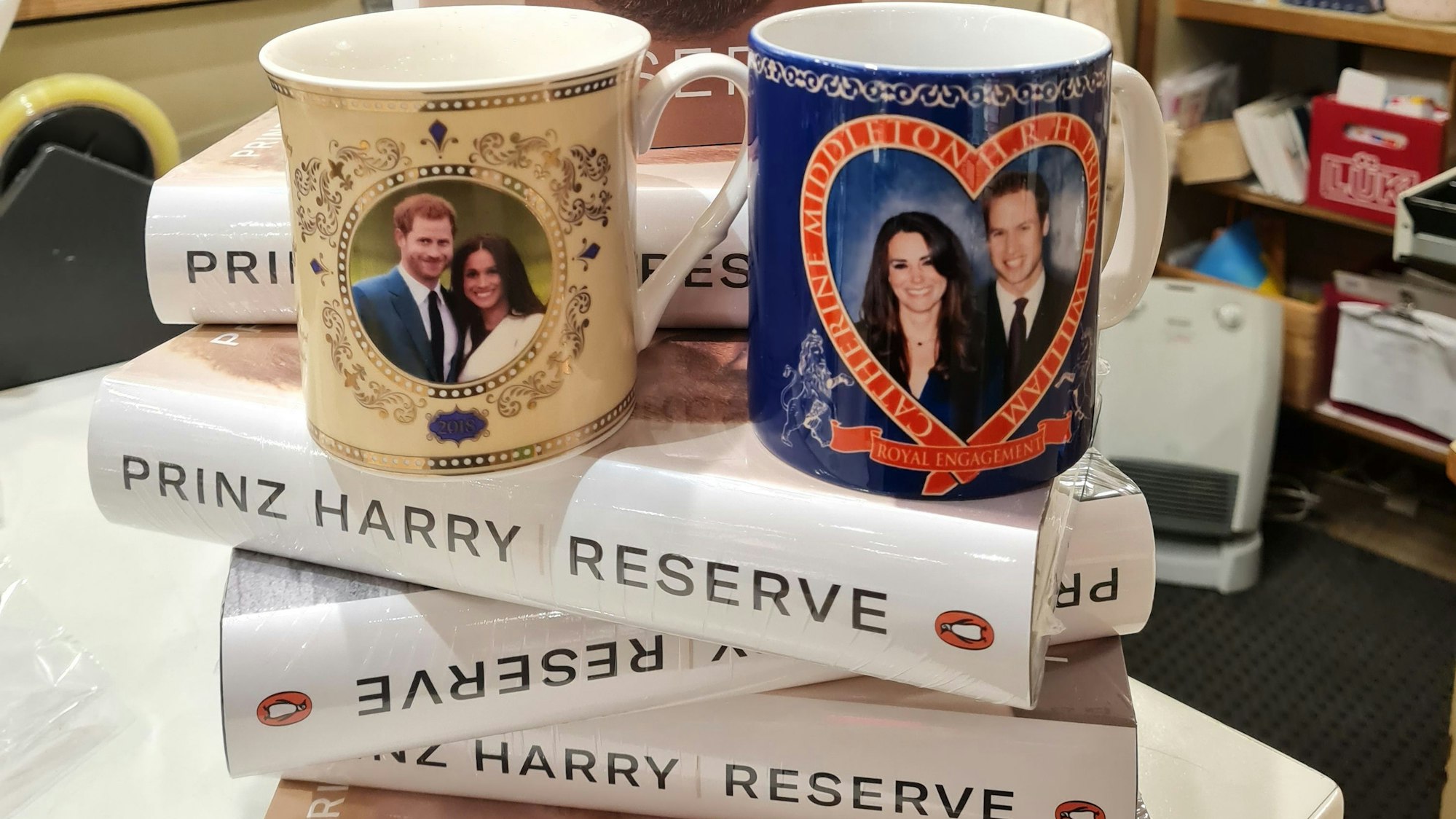 Auf einem kleinen Stapel des Buches von Prinz Harry steht eine Tasse mit dem Foto von Harry und Meghan und eine mit dem Foto von William und Kate.